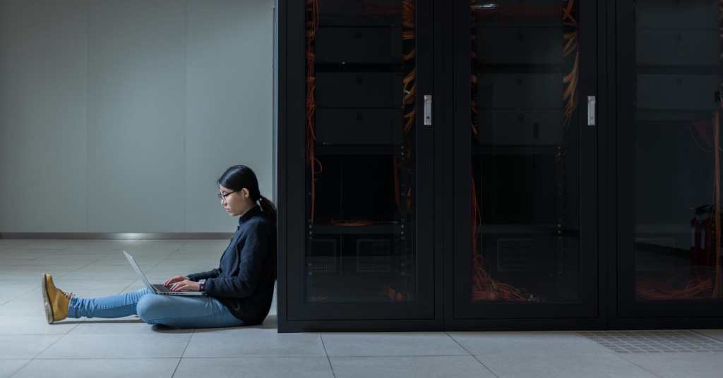 Woman sitting on the floor, working on her laptop while leaning against a bank of servers