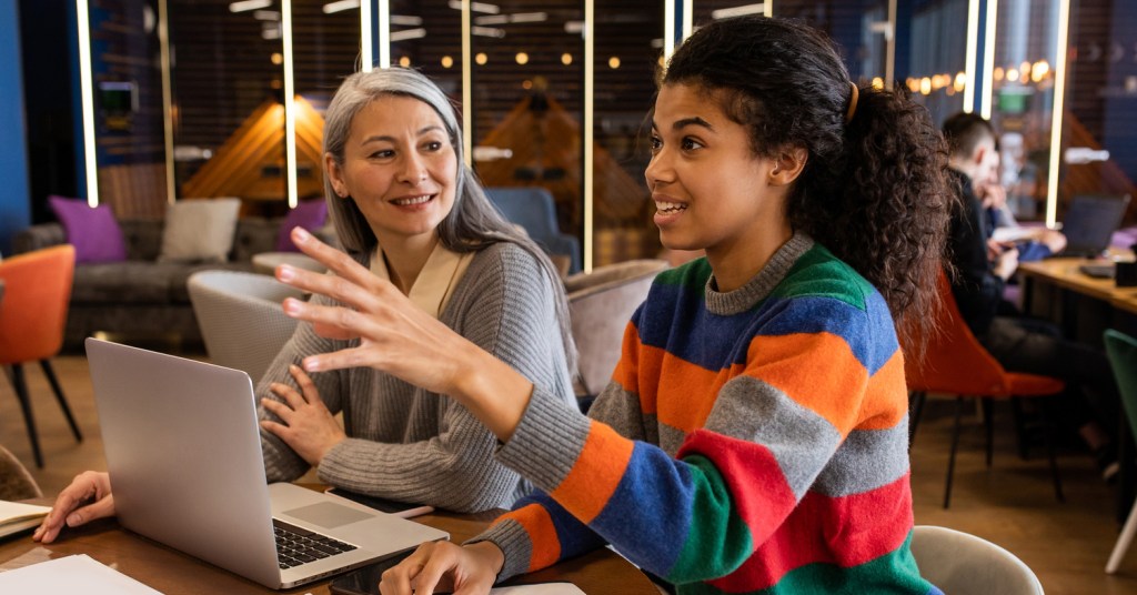 Two long haired people sitting next to each other, one colorfully dressed looks ahead while gesturing with their hand while the other person looks towards their companion a laptop is placed in front of the two of them
