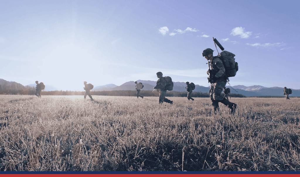 Soldiers with packs walking across a field with mountains in the background.